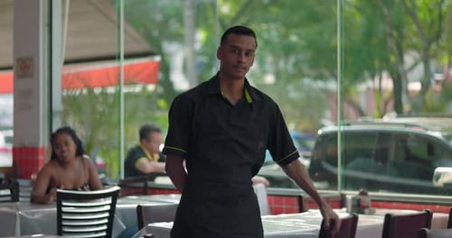 Young restaurant staff member posing near table and glass window, leaning on chair with neutral