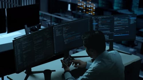 Hombre asiático trabajando con un teléfono móvil mientras escribe código con múltiples pantallas de ordenador en la oficina