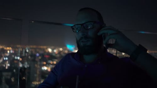 Young man chatting on cellphone and enjoying cocktail at rooftop bar at night
