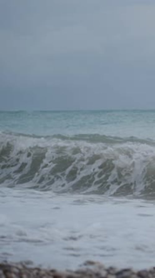 Vertical video. Stormy Sea View from the Pebble Beach, Dark Blue Tones, and Strong Waves Crashing
