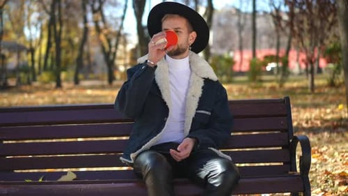 Handsome Young Man Sitting on Bench in Autumn Park Drinking Coffee Looking Away Waving
