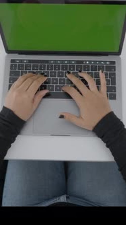 A Young Woman Dressed in Black is Sitting at a Clean Desk Watching a Laptop with a Green Screen