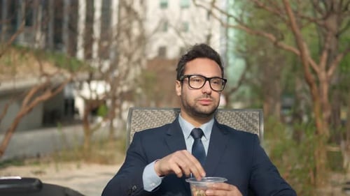 Pensive millennial businessman eating nuts in park outdoors enjoying lunch break. Thoughtful man in