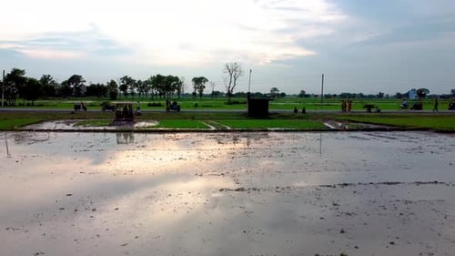 Silhouetted people on a roadside watch a tractor plow green muddy field in the country side for cult