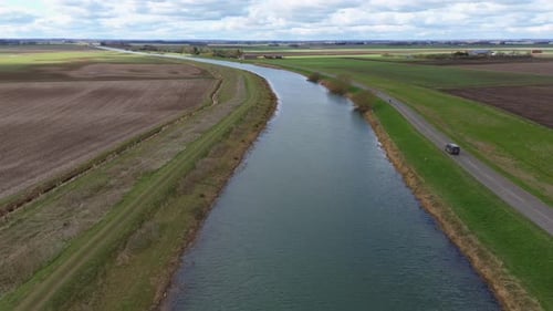 A long, narrow river runs parallel to a countryside road in the flat farmlands of Lincolnshire, UK.