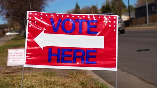 Vote Here Sign Pointing Left with People Driving Cars in Background, Close Up