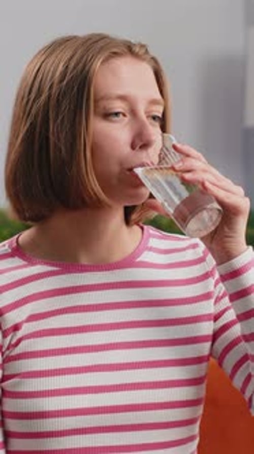 Durstige junge Frau sitzt zu Hause und hält ein Glas natürlichen Aquas in der Hand, macht Schlucke und trinkt stilles Wasser