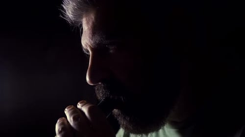 Close up of bearded men inhaling e-cigarette device at night in front of light 01.