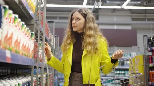Thoughtful Shopper Woman in a Yellow Jacket Browses Products in a Grocery Store Aisle with a