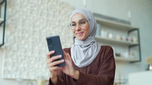 Lächelnde Brünette mit Brille hält ihr Smartphone in der Hand, scrollt und schaut soziale Medien bei Light Domestic