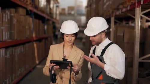 Young man in work uniform and woman in business suit working in warehouse with boxes