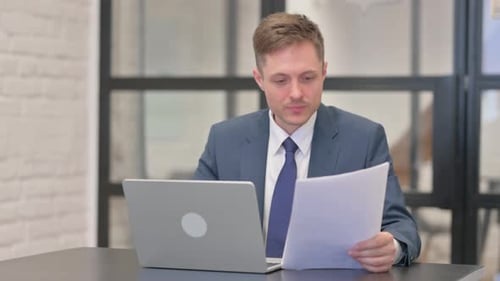 Man Looks at Laptop and Paperwork in Office