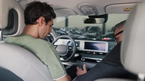 A Brunette Guy in a Green Tshirt Sits in a Car with a Manager in a Business Suit at a Car Dealership