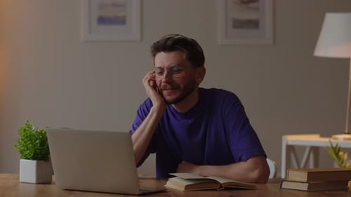 A Man with Glasses Sits at a Table with Books and Talks to Someone Through a Laptop Webcam A College