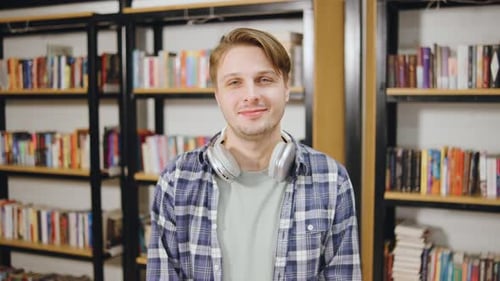 Young Man Stands in Front of Bookshelves with Headphones Around His Neck in a Library Setting During