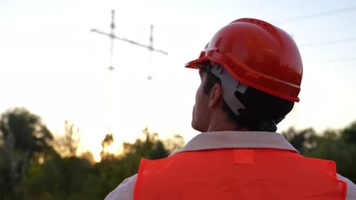 Young Engineer Standing Outdoors While Looking at the High Voltage Power Lines Back View Energy