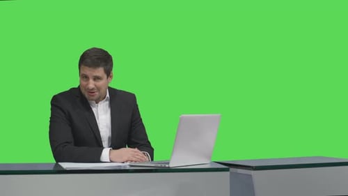 Media broadcaster is sitting at a table and talking on a mock-up green screen in the background.