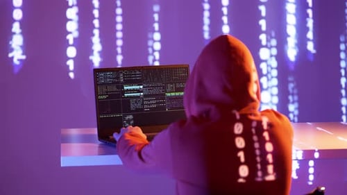 Un hacker cagoulé tape sur un ordinateur portable entouré de code binaire projeté et d'écrans de terminal. La scène