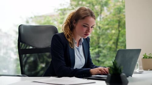 in a Modern Office with a Large Window a Beautiful Worker in a Blue Shirt with a Laptop Looks at the