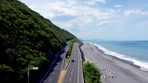 Coastal road curving along a lush hillside and beach, blue sea on a sunny day, aerial shot