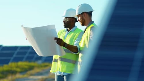 Shot of Two Engineers with Project Plan on the Solar Farm in Special Uniform Standing Discuss the