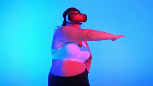 Woman with overweight in a tracksuit and VR headset posing in a studio with blue background and red