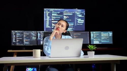 Female Programmer Thinking About Something While Writing Code By A Laptop Using Multiple Monitors