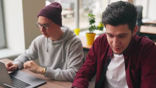 Zwei Hipster-Mitarbeiter tauschen Ideen zum Projekt aus und sitzen mit Laptops im geräumigen Großraumbüro