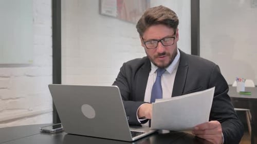 Man Reviews Documents at Laptop in Office
