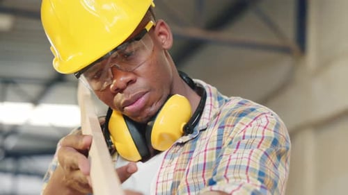 African American Carpenter Inspecting a Piece of Wood While Carrying a Wooden Plank in a Workshop
