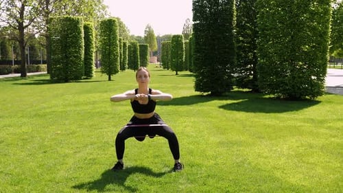 A Woman Trains Her Legs with Squats Using Rubber Bands for Fitness in a Park