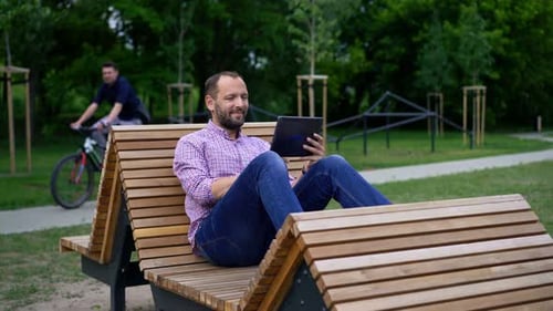 Man Browsing Internet On Tablet Sitting On Bench In City Park