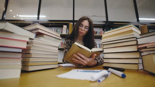 Student Reading and Taking Notes at a Desk Surrounded By a Stack of Books in a Library During