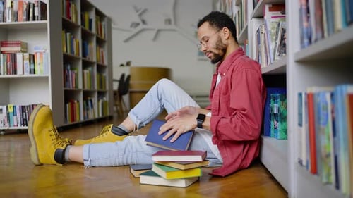 Multiethnic Postgraduate Male Student Leafs Through the Book Pages Sitting on the Library Floor