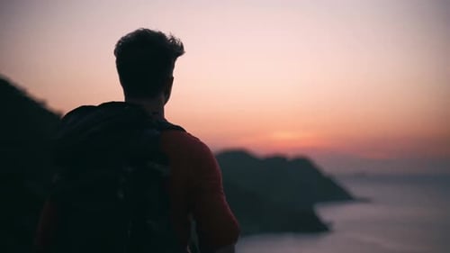 A young boy with backpack standing alone watching sunset along the mountain and sea.