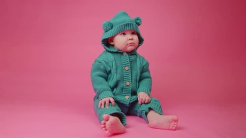Portrait of a Cute Baby in a Warm Knitted Jumpsuit with a Hood on a Pink Background