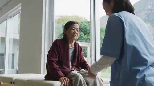 Smiling asian female physiotherapist treating knee of senior female patient and talking at surgery