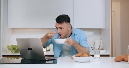 Man sitting at kitchen table eating cereal for breakfast and watching funny videos on laptop