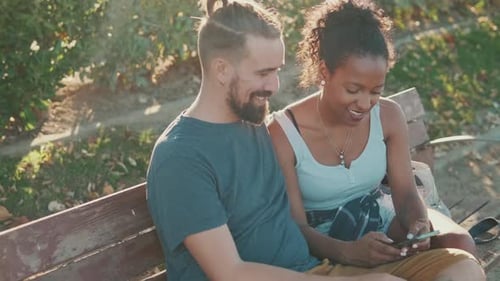 Happy smiling interracial couple watching video on mobile phone while sitting on bench