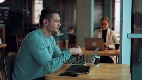 Tired man sitting in front of laptop rubs his eyes holding glasses in his hand.