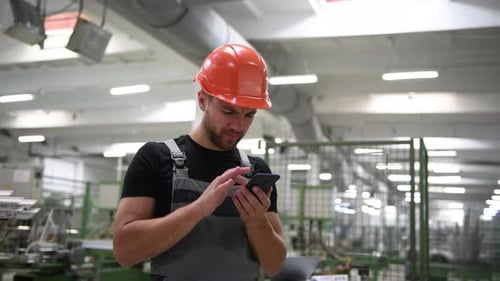 Man in uniform and hard hat stands in the factory and uses phone