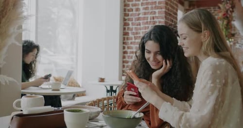 Joyful Young Women Looking at Smartphone Screen Talking Sharing Content Sitting at Table in Cafe