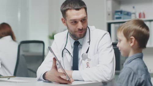 In Hospital. Handsome Smiling Doctor Shows Something on a Tablet Computer to a Smiling Little Boy a