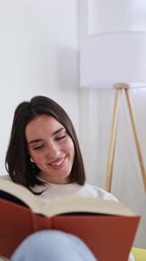 Young Woman Reading Book and Smiling While Relaxing in Bright Living Room
