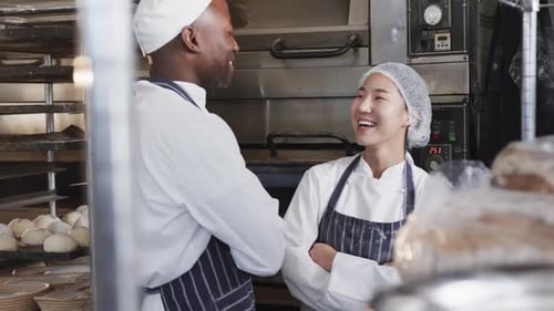 Happy diverse bakers in bakery kitchen with arms crossed talking in slow motion