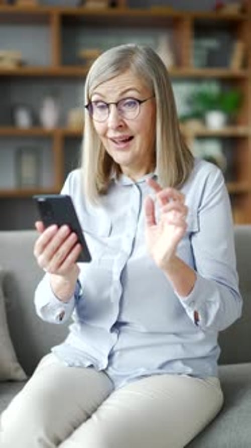 Vertical video. Happy senior woman with gray hair received great news on smartphone sitting on sofa