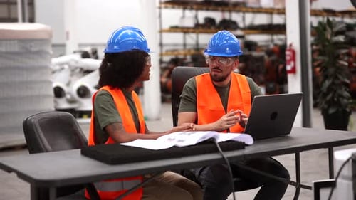 Zwei gemischtrassige Ingenieure, die Sicherheitswesten und Helme tragen, benutzen einen Laptop und besprechen ein Projekt