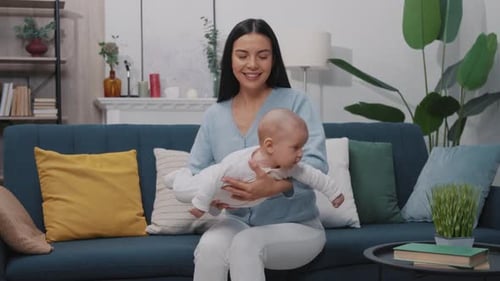 Lustige glückliche junge Mutter spielt mit ihrem neugeborenen Baby, hält sie fest und sitzt auf einer Couch in einem