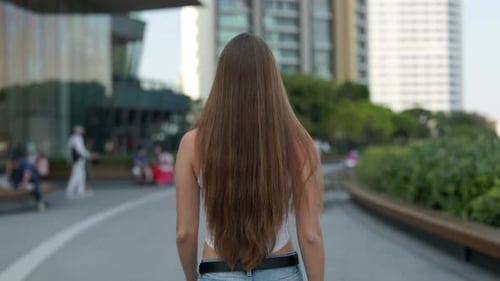 Young Woman with Long Brown Hair Walks Along City Center Street Backside View