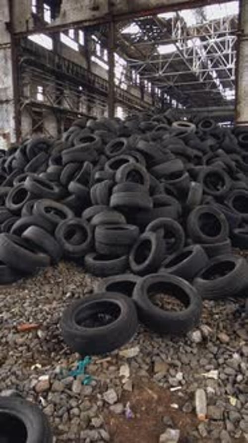A bunch of car tires are in an abandoned high hangar with holes in the windows of building.
